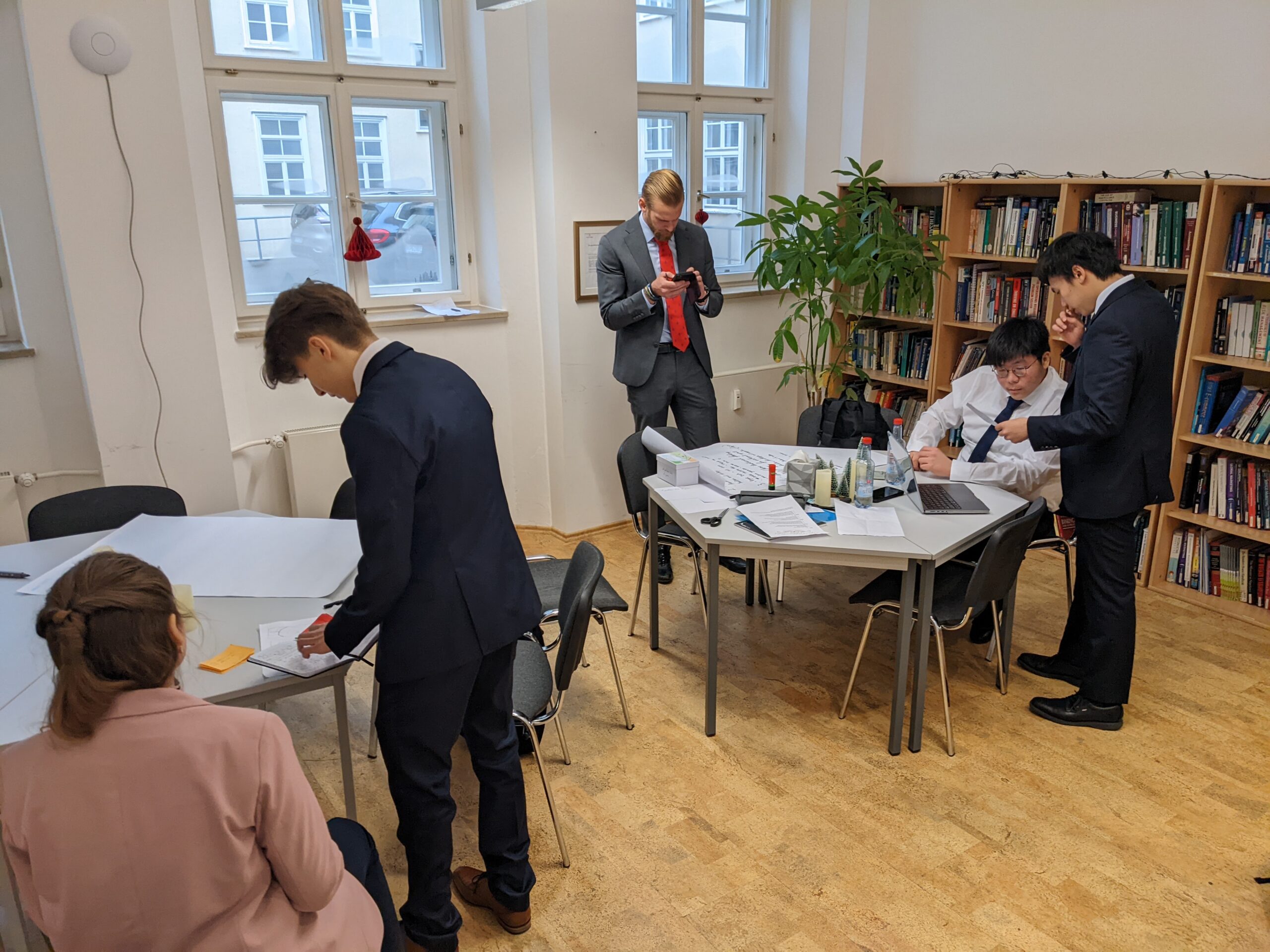 The setting of this photo is the library at Globe Business College Munich. There are two tables on a cork floor. Five students are in two groups: one of two students, one of three. THey're centered around paper and a laptop to finish their final projects.