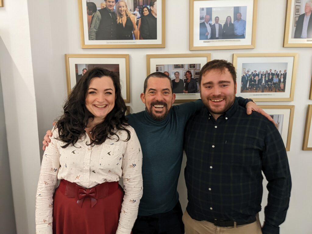Two teachers from Cardiff Sixth Form in the UK stand in front of a wall of testimonials and photos at Globe Business College Munich with Packy Lee, actor from Peaky Blinders.