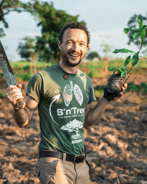 Chris Kaiser stands in a field wearing a green shirt. The print on the shirt is a tree with an arrow up to a pair of human lungs and 02 (for oxygen) above the arrow. The shirt also features a slogan. Chris is holding a machete in one hand a tree sapling in the other as he plants trees in a remote Asian field